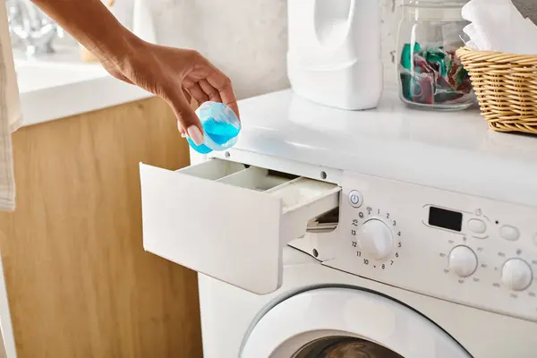 Una mujer afroamericana sostiene una cápsula de gel frente a una lavadora mientras lava la ropa en un baño. - foto de stock