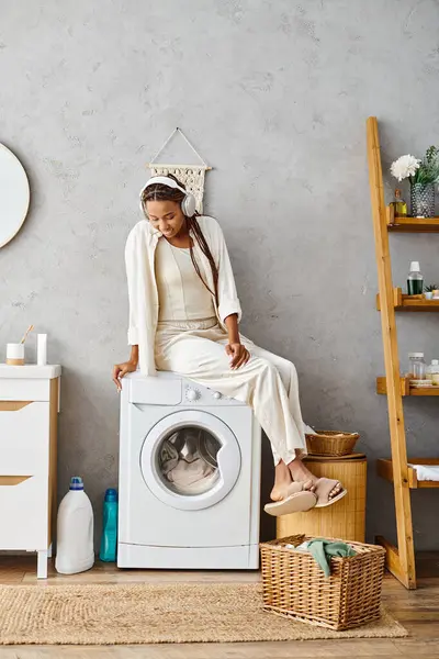 Eine afroamerikanische Frau mit Afro-Zöpfen sitzt stolz auf einer Waschmaschine und kümmert sich um die Wäsche in ihrem Badezimmer. — Stockfoto