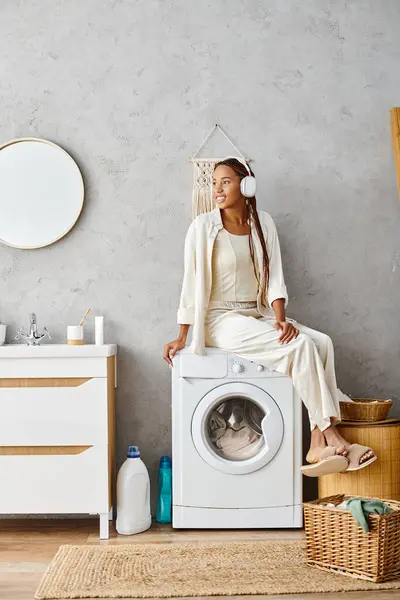 Eine afroamerikanische Frau mit Afro-Zöpfen sitzt auf einer Waschmaschine und macht einen Moment der Ruhe während ihrer Wäsche-Routine. — Stockfoto