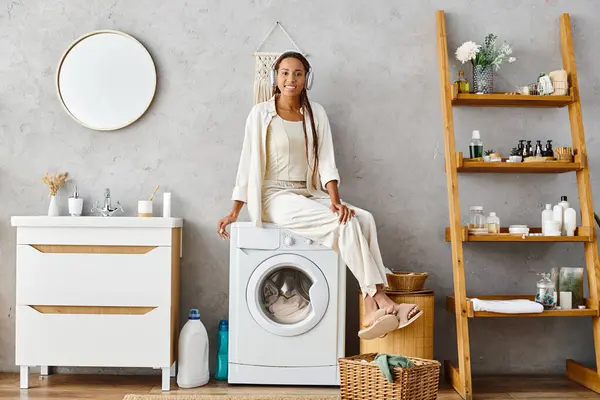 Una mujer afroamericana con trenzas afro se sienta con confianza en una lavadora haciendo la colada en un baño. - foto de stock
