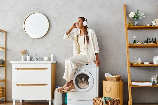 Femme afro-américaine avec des tresses afro faire la lessive, assis au sommet d'une machine à laver dans une salle de bain. — Photo de stock
