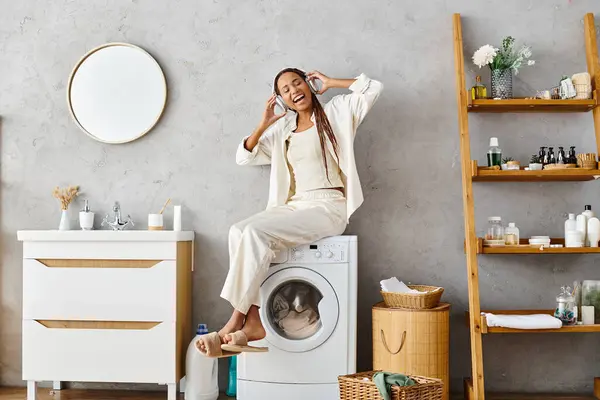Africano mulher americana com tranças afro confortavelmente sentado no topo de uma máquina de lavar roupa enquanto fazendo a roupa em um banheiro. — Fotografia de Stock