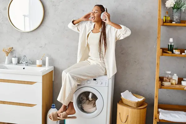 Mulher afro-americana com tranças afro senta-se calmamente em uma máquina de lavar roupa, lavando roupa em um banheiro. — Fotografia de Stock