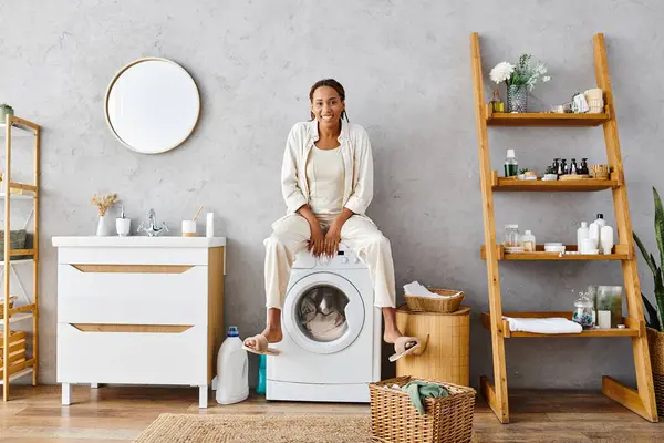 Una mujer afroamericana con trenzas afro sentada encima de una lavadora, lavando ropa en un baño. - foto de stock