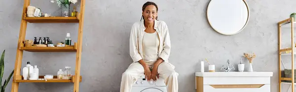 Una mujer afroamericana con trenzas afro se sienta encima de una lavadora haciendo la colada en un acogedor entorno de baño. - foto de stock