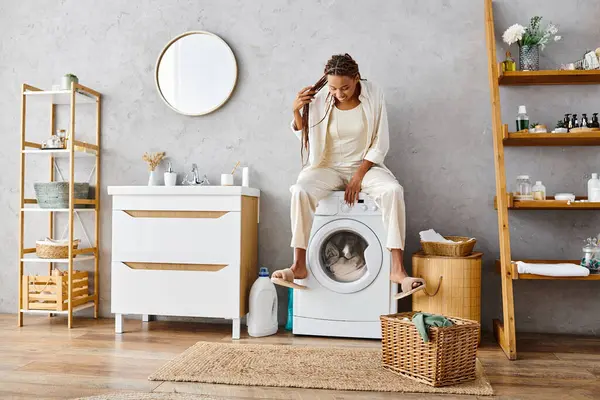 Femme afro-américaine avec des tresses afro se trouve au sommet d'une machine à laver, faire la lessive dans une salle de bain. — Photo de stock