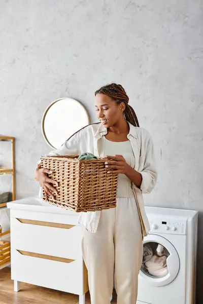 Femme afro-américaine avec des tresses tenant un panier en osier devant une machine à laver dans une salle de bain. — Photo de stock