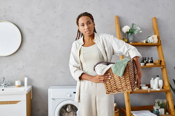 Una mujer afroamericana con trenzas afro lavando ropa en un baño sereno, sosteniendo una cesta. - foto de stock