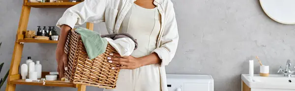 Une femme d'ascendance africaine faisant la lessive, tenant un panier dans un cadre de salle de bain serein. — Photo de stock