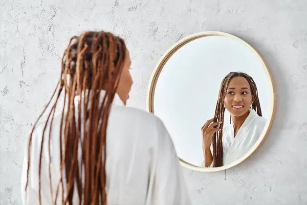 Une Afro-Américaine avec des tresses afro se brosse les cheveux devant un miroir, baignée dans une douce lumière de salle de bain. — Photo de stock