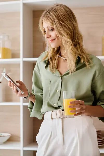 Una donna in piedi in una cucina con un cellulare in una mano e un bicchiere di succo d'arancia nell'altra. — Foto stock
