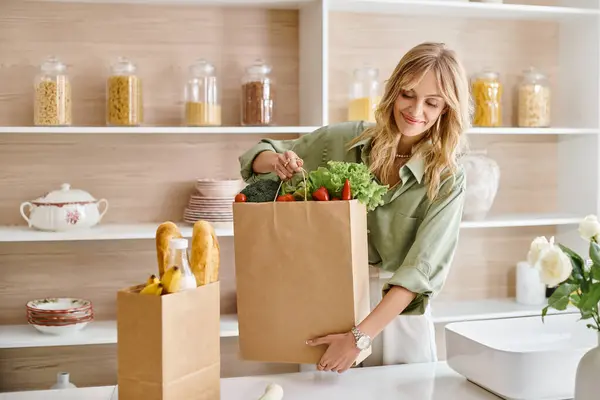 A woman in a kitchen holding a grocery bag filled with fresh vegetables. — Stock Photo