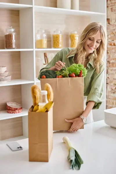 Mujer de pie en la cocina, sosteniendo una bolsa llena de verduras frescas. - foto de stock