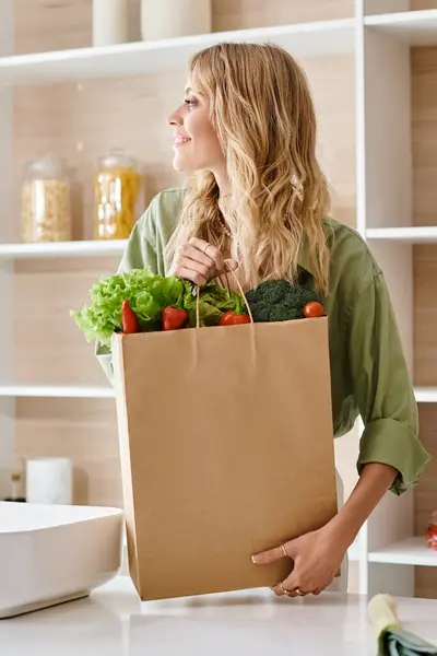 A woman holding a grocery bag filled with fresh vegetables in a kitchen. — Stock Photo