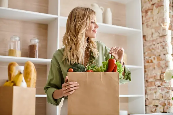 Una mujer de pie en una cocina, sosteniendo una bolsa llena de verduras. - foto de stock