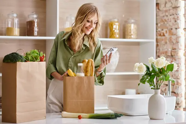 Woman in kitchen looks at phone with bag of food. — Stock Photo