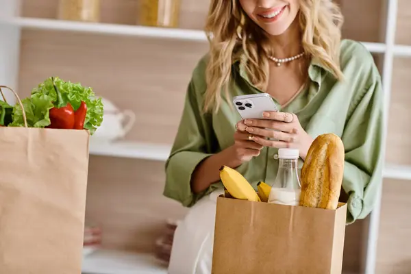 Жінка на кухні дивиться на свій мобільний телефон, тримаючи мішок їжі. — стокове фото