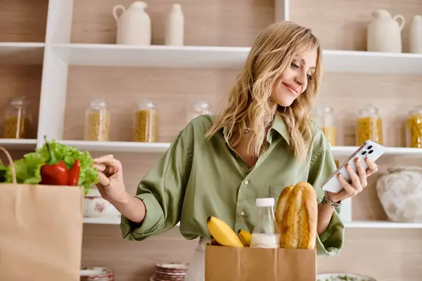 Una mujer con una camisa verde de pie en una cocina, sosteniendo un teléfono celular y una bolsa de comida. - foto de stock