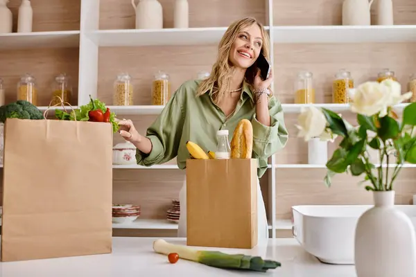 Mujer multitarea con teléfono celular y bolsa de compras en la cocina. - foto de stock