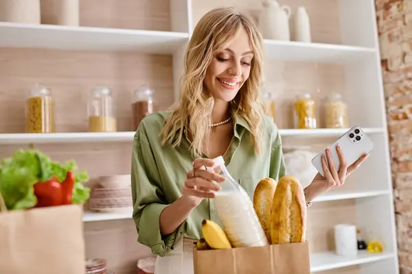 Una mujer parada en una cocina sosteniendo una bolsa de pan y un teléfono celular. - foto de stock