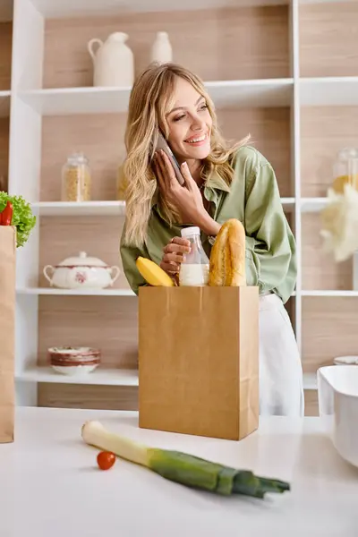 Una mujer en una cocina de apartamento hablando por un teléfono celular mientras sostiene una bolsa de compras. - foto de stock