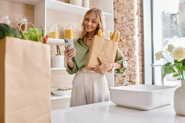 A woman holding a paper bag and a cell phone in a kitchen. — Stock Photo