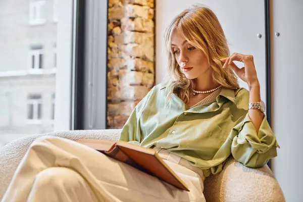 Una mujer sentada en una silla, absorta en un libro en casa. - foto de stock