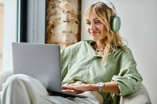 Eine Frau sitzt auf einer Couch, trägt Kopfhörer und benutzt einen Laptop in einer gemütlichen Wohnung. — Stockfoto