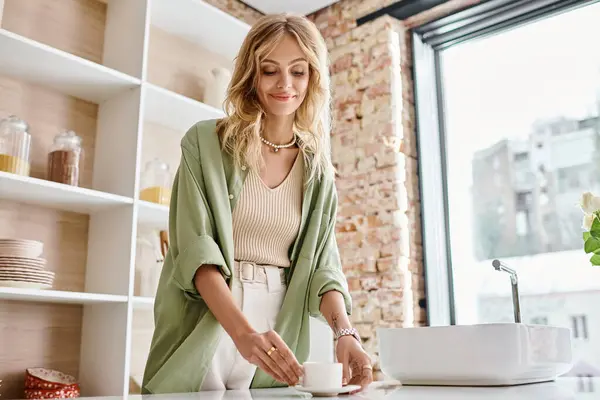Happy young blonde woman in green cardigan smiling and taking cup of coffee in kitchen — Stock Photo