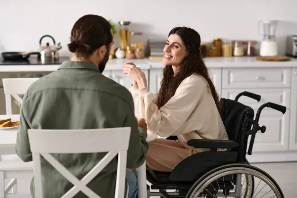 Бородатый веселый мужчина наслаждается завтраком со своей веселой женой-инвалидом в инвалидном кресле за завтраком — стоковое фото