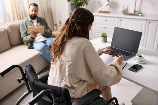Привлекательная женщина-инвалид в инвалидной коляске работает из дома на ноутбуке рядом со своим мужем-читателем — стоковое фото