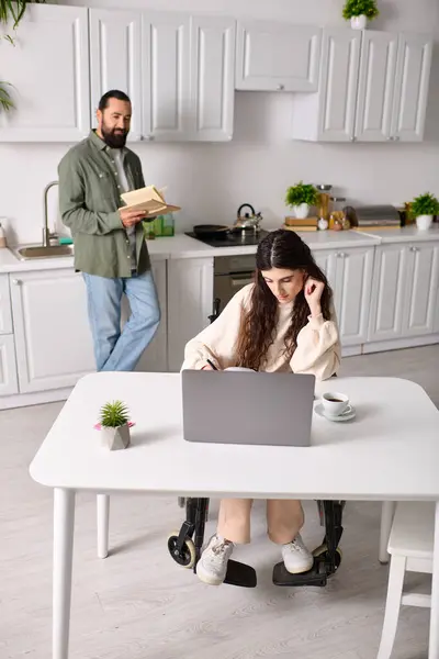 Behinderte Frau im Rollstuhl arbeitet von zu Hause aus am Laptop neben ihrem lesenden Mann — Stockfoto