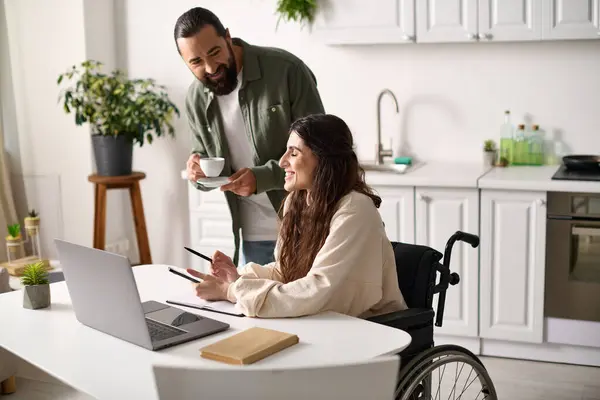 Atractivo hombre bebiendo café junto a su alegre esposa discapacitada en silla de ruedas trabajando en el portátil - foto de stock