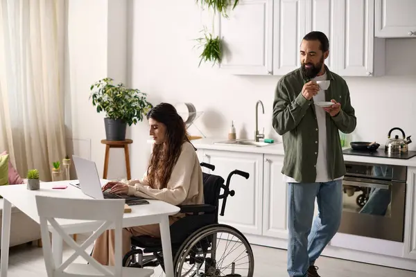 Красивый мужчина пьет кофе рядом со своей веселой женой-инвалидом в инвалидном кресле, работающей за ноутбуком — стоковое фото