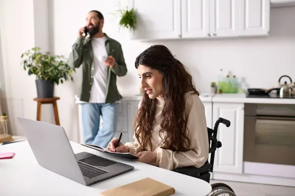 Веселая женщина-инвалид в инвалидной коляске работает удаленно рядом с мужем разговаривая по телефону на заднем плане — стоковое фото