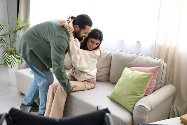 Bearded man in homewear helping his wife with mobility disability to get to couch from wheelchair — Stock Photo