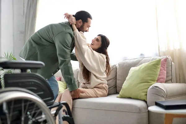 Amoroso hombre en ropa de casa ayudar a su esposa con discapacidad de movilidad para llegar al sofá de la silla de ruedas - foto de stock