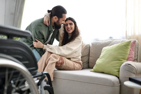 Amoroso hombre en ropa de casa ayudar a su esposa con discapacidad de movilidad para llegar al sofá de la silla de ruedas - foto de stock