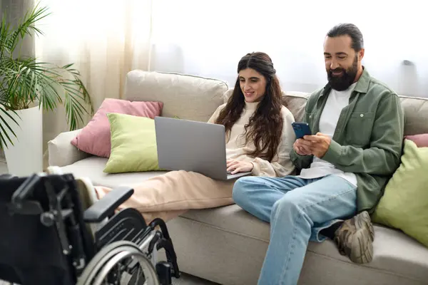 Bella donna con disabilità motoria seduta sul divano con computer portatile accanto al marito con telefono — Foto stock