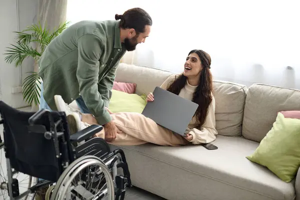 Donna attraente con disabilità motoria seduta sul divano con computer portatile accanto al marito amorevole — Foto stock