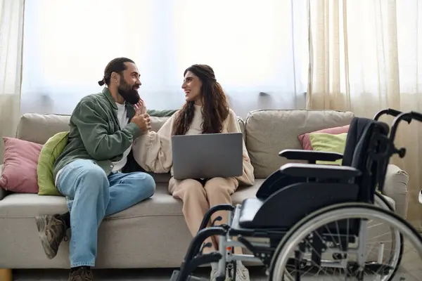 Alegre hombre mirando a la computadora portátil al lado de su discapacitada hermosa esposa alegre mientras está sentado en el sofá en casa - foto de stock