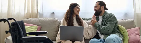 Freudiger Mann, der neben seiner behinderten fröhlichen schönen Frau auf dem Sofa zu Hause auf den Laptop schaut — Stockfoto