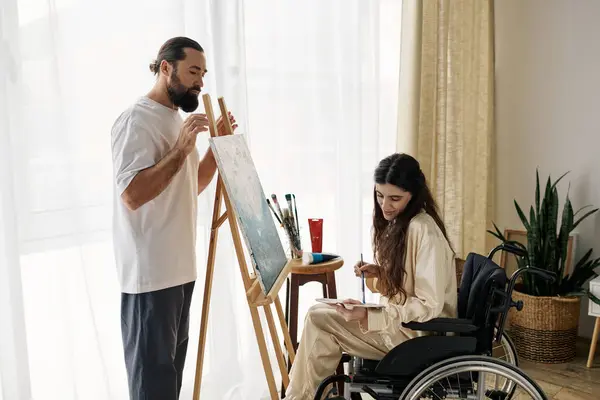 Bell'uomo barbuto guardando la sua bella moglie disabile sulla sedia a rotelle che dipinge sul cavalletto a casa — Foto stock