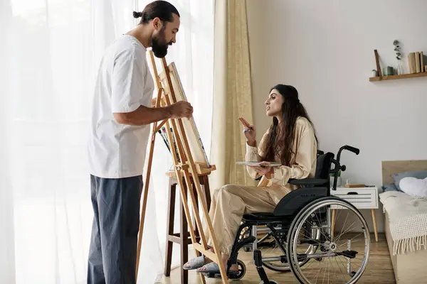 Guapo barbudo hombre viendo su discapacitado hermosa esposa en silla de ruedas pintura en caballete en casa - foto de stock