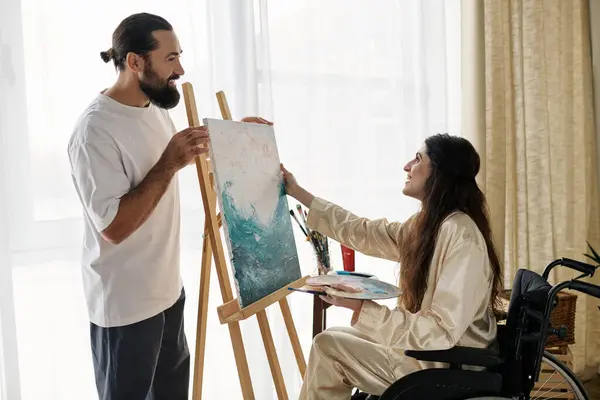 Bell'uomo barbuto guardando la sua bella moglie disabile sulla sedia a rotelle che dipinge sul cavalletto a casa — Foto stock