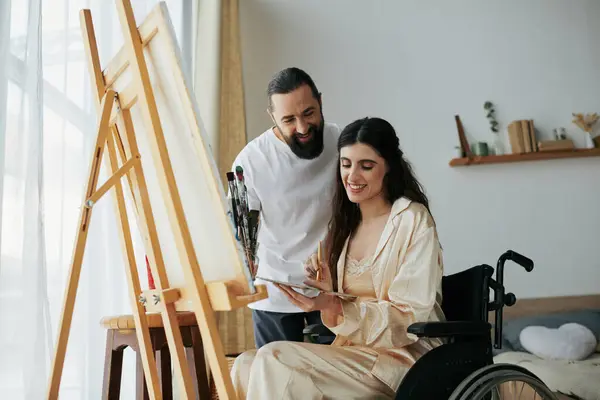 Atractivo barbudo hombre viendo su discapacitado hermosa esposa en silla de ruedas pintura en caballete en casa - foto de stock