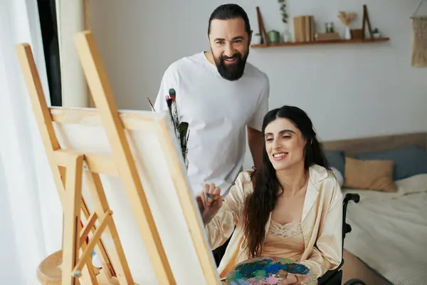 Attraktiver bärtiger Mann, der seiner behinderten schönen Frau beim Malen im Rollstuhl auf der Staffelei zu Hause zusieht — Stockfoto