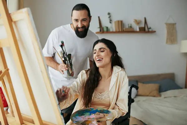 Amorevole marito allegro aiutando la sua moglie inclusiva di bell'aspetto a dipingere sul cavalletto mentre a casa — Foto stock