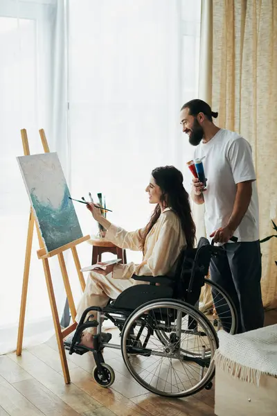 Gut aussehender bärtiger Mann beobachtet behinderte schöne Frau auf Rollstuhl-Malerei auf Staffelei zu Hause — Stockfoto