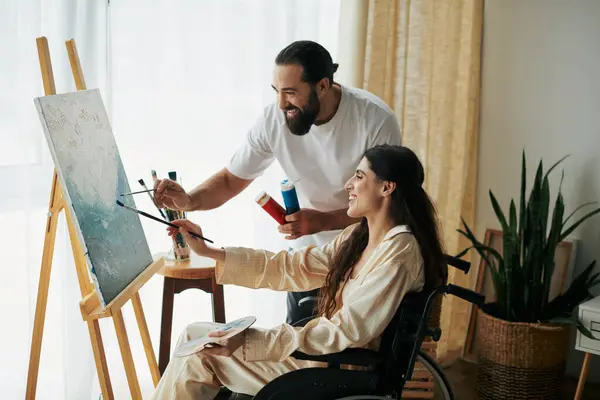 Gut aussehender bärtiger Mann beobachtet behinderte schöne Frau auf Rollstuhl-Malerei auf Staffelei zu Hause — Stockfoto